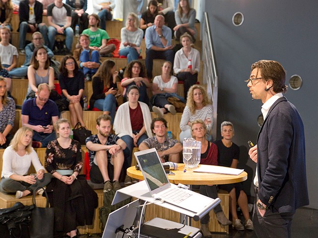 Een man achter een tafel met microfoon en praat voor studenten