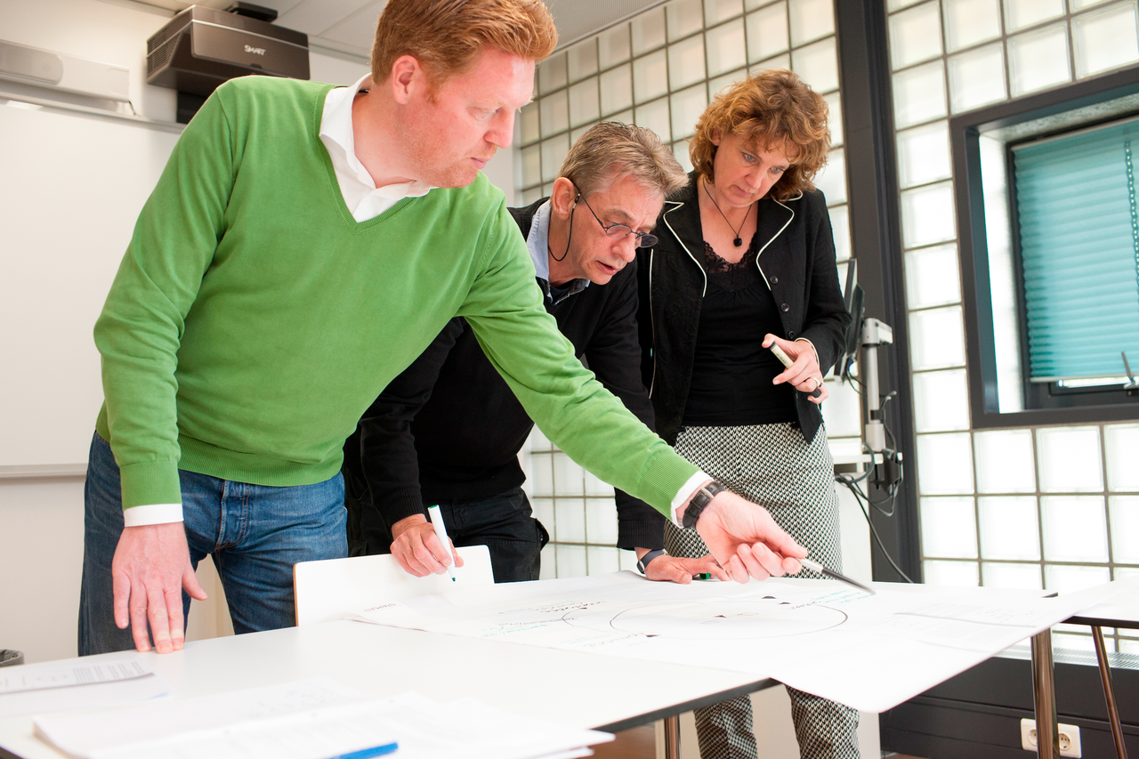 Drie mensen staan boven tafel met papieren en overleggen