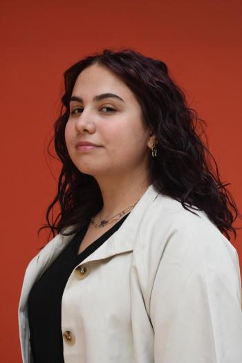 Portret van een vrouw met donker, krullend haar in een lichte jas en zwart shirt, tegen een oranje achtergrond.