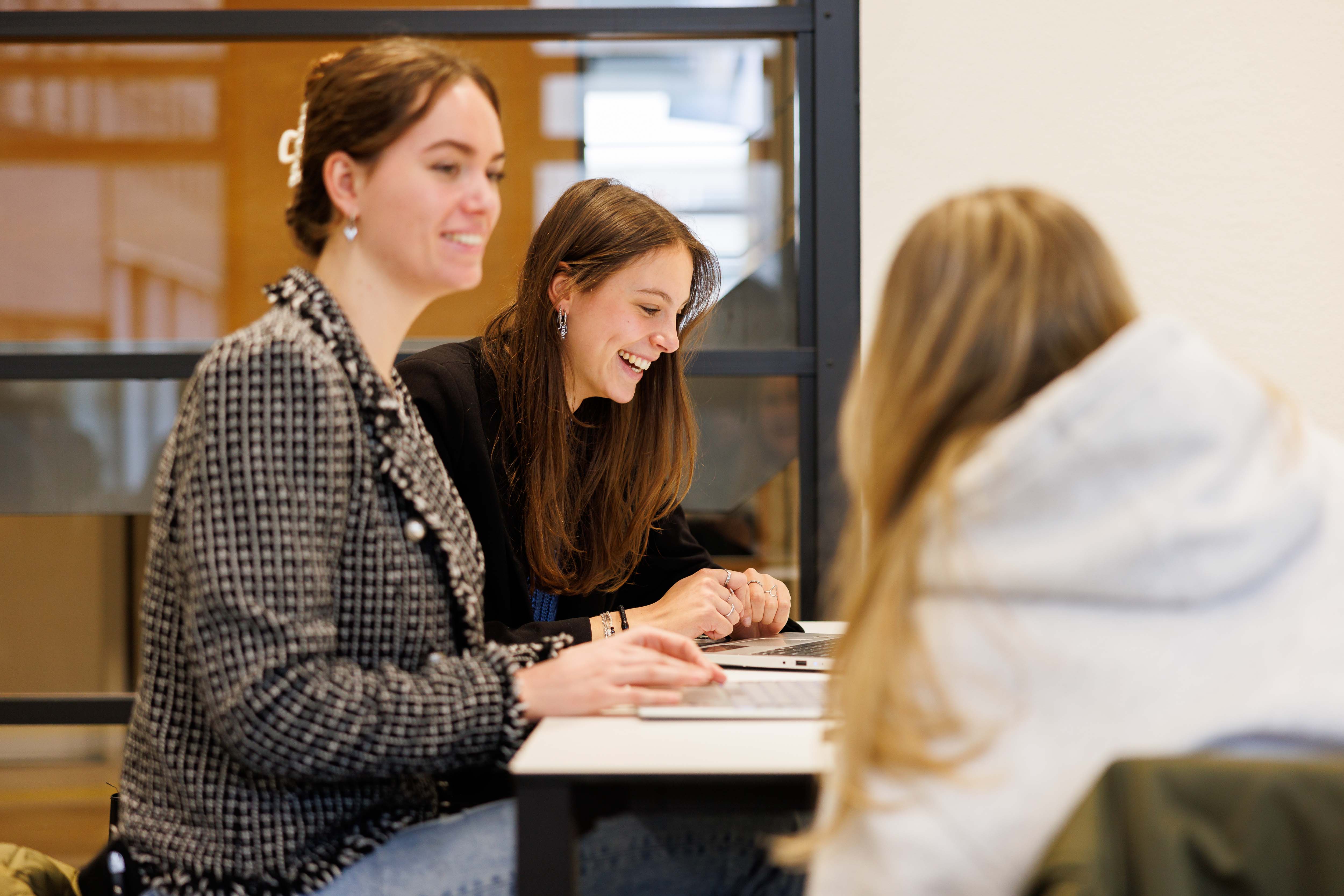 Drie jonge vrouwen werken samen aan een tafel met laptops, lachend en geconcentreerd.