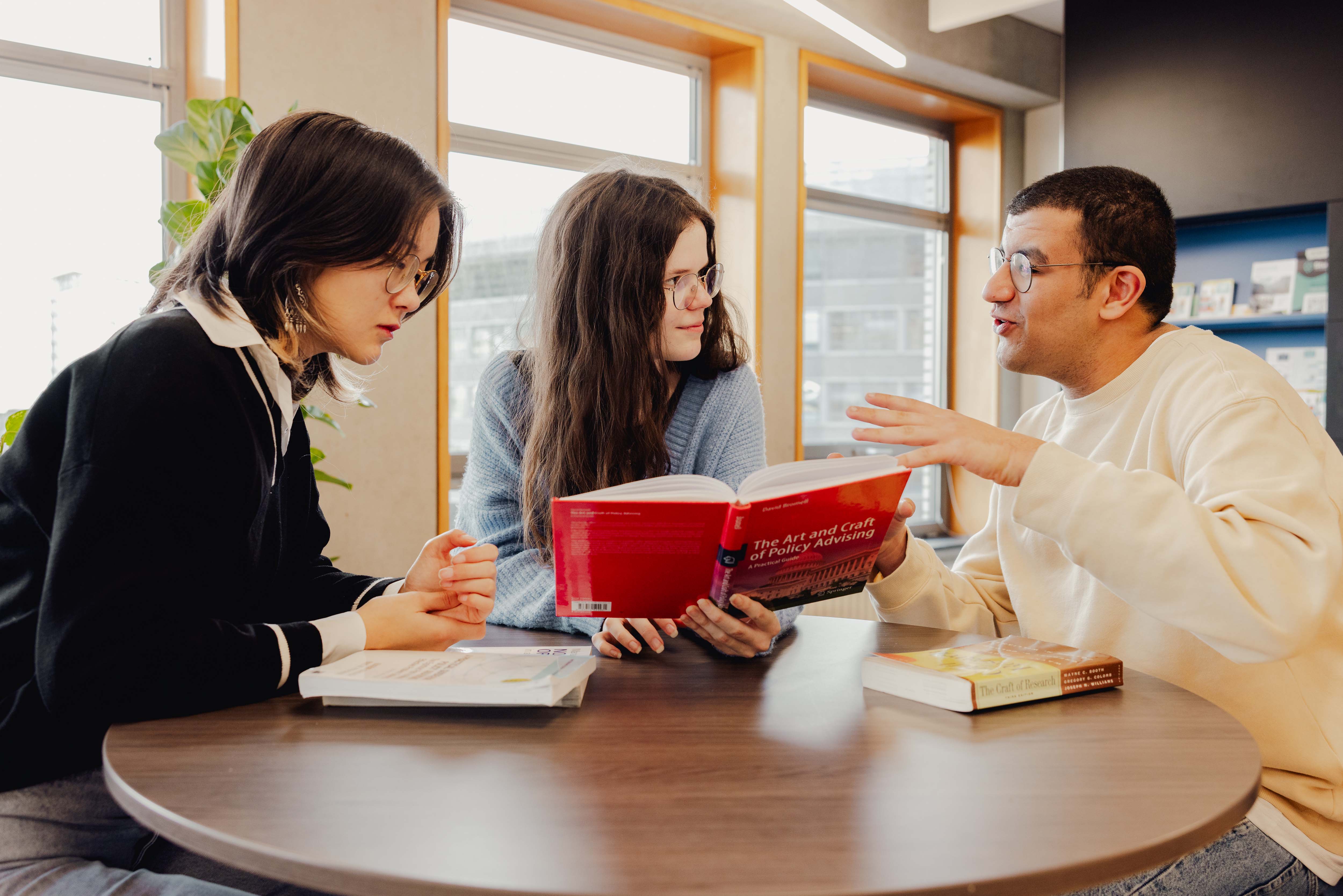 Drie studenten lezen en bespreken samen een boek aan een tafel in een lichte kamer.