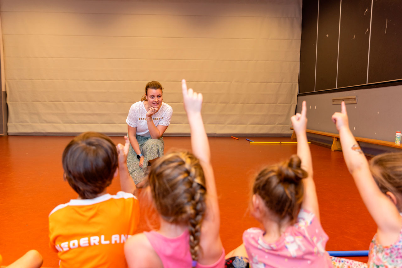 Vrouw praat met groep kinderen, waarvan er meerdere hun hand opsteken.