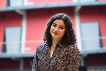 Portret van een vrouw met krullend zwart haar, gekleed in een donkerblouse met patroon, staand voor een rood gebouw.