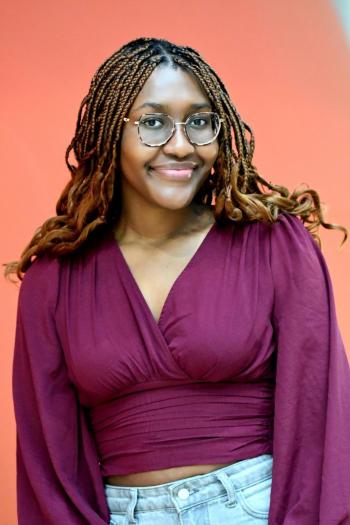 Portret van een vrouw met gevlochten haar en bril, in een bordeauxrode top en spijkerbroek, voor een oranje achtergrond.