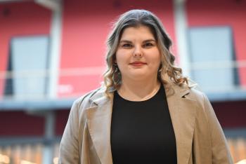 Portret van een vrouw met halflang, golvend haar, in een beige jas en zwart shirt, staand voor een rood gebouw.