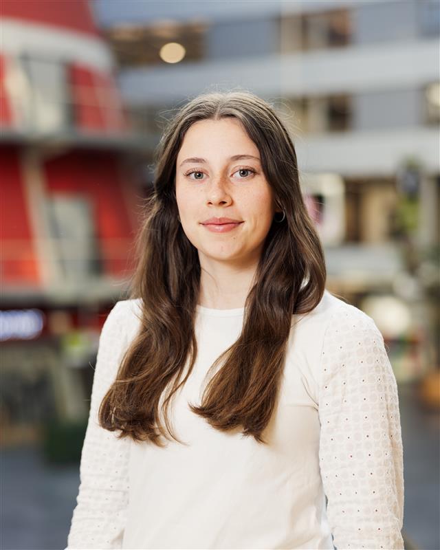 Jonge vrouw met lang bruin haar, gekleed in een witte blouse, kijkt rustig in de camera.