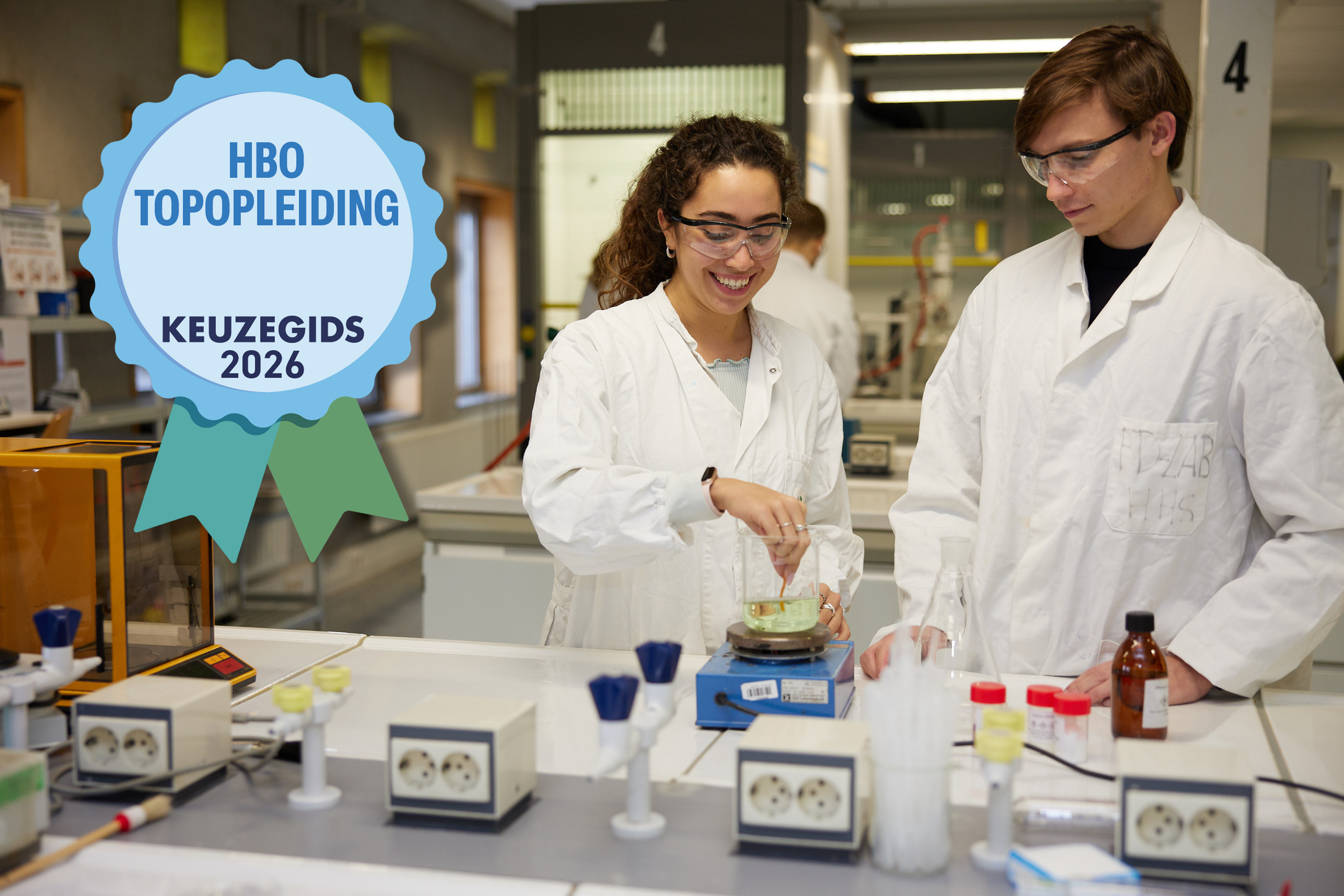 Twee studenten in labjassen werken samen in een laboratorium; logo "HBO Topopleiding Keuzegids 2026" staat op de voorgrond.