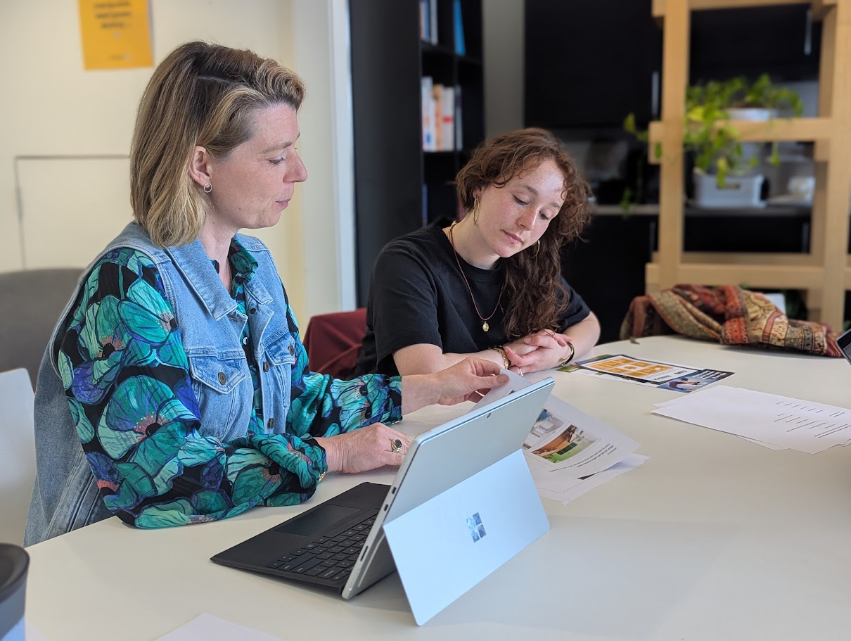 Twee collega’s zitten aan een vergadertafel en bekijken samen documenten en een tablet tijdens een overleg op kantoor.