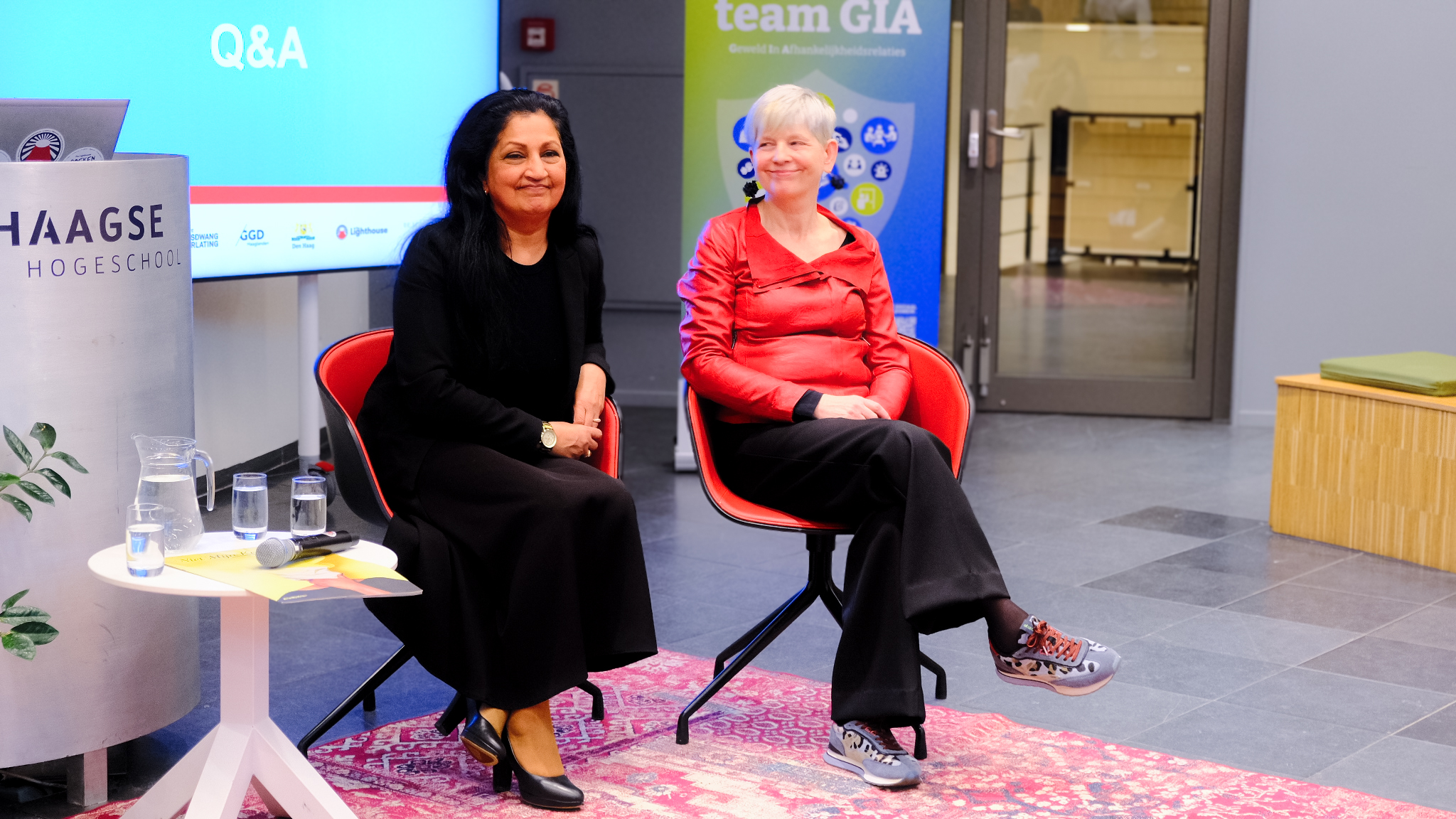 Two speakers seated on stage during a Q&A event at a university, with a screen, podium, and microphone visible