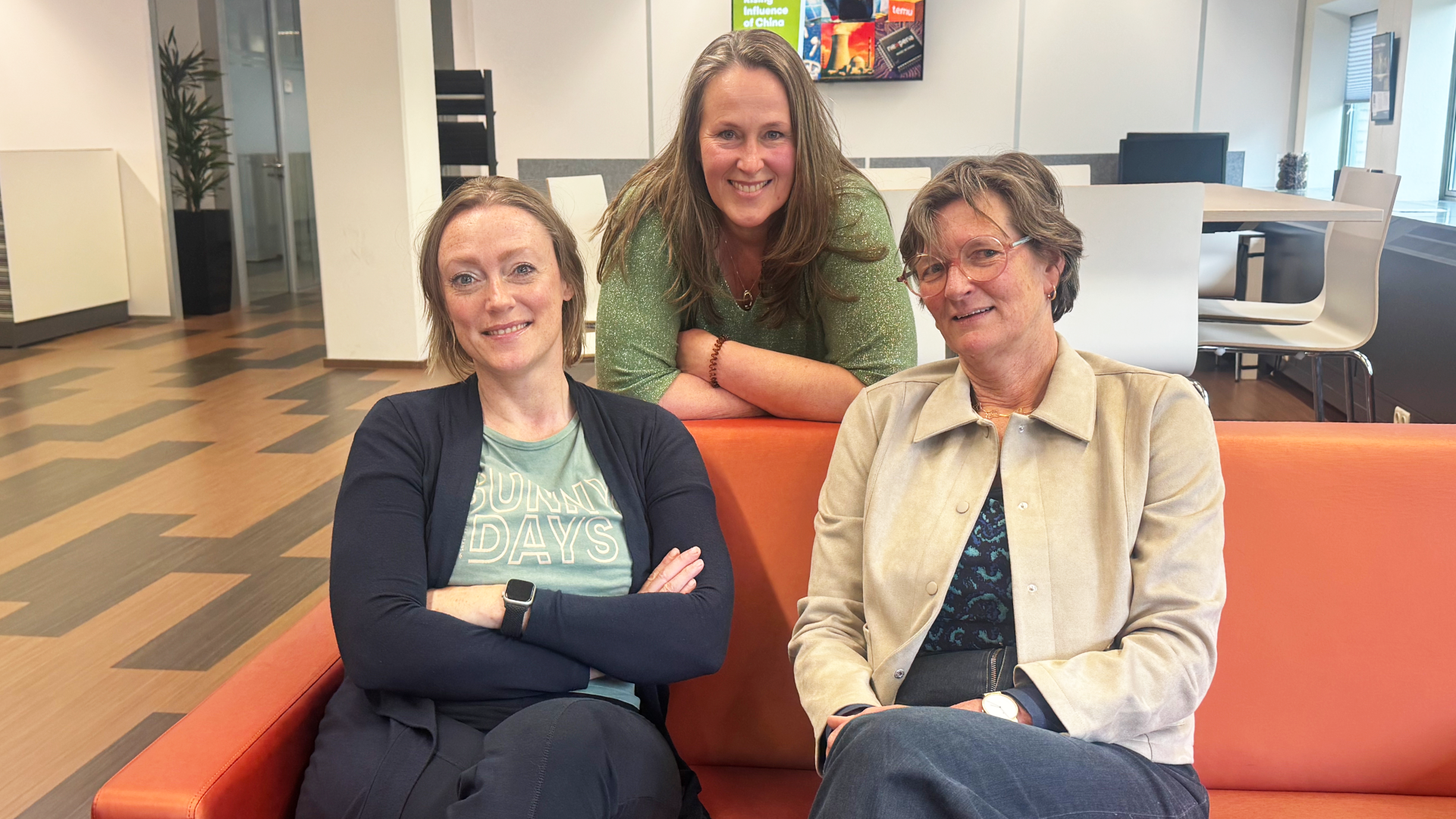 Drie personen op en achter een oranje bank in een lichte kantoorruimte.