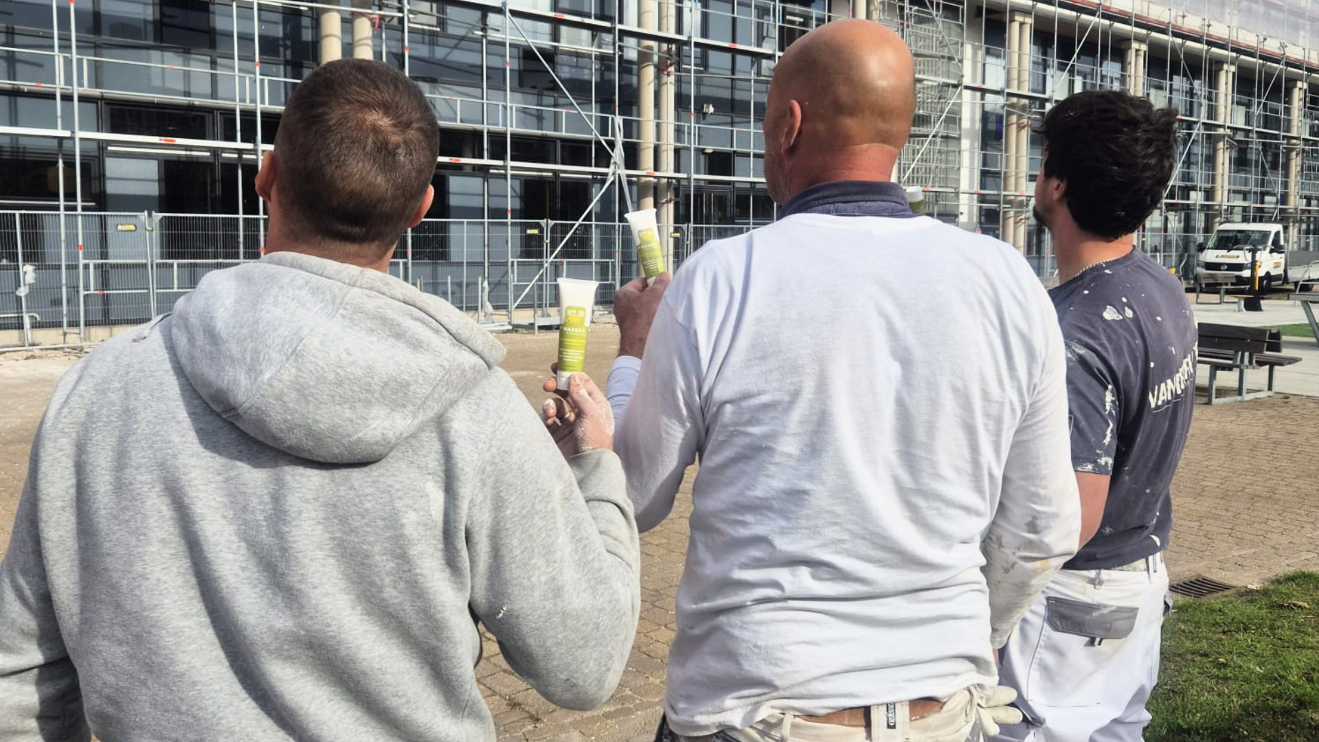 Drie bouwmedewerkers bekijken een gebouw in aanbouw met steigers en houden tubes met bouwmateriaal vast.
