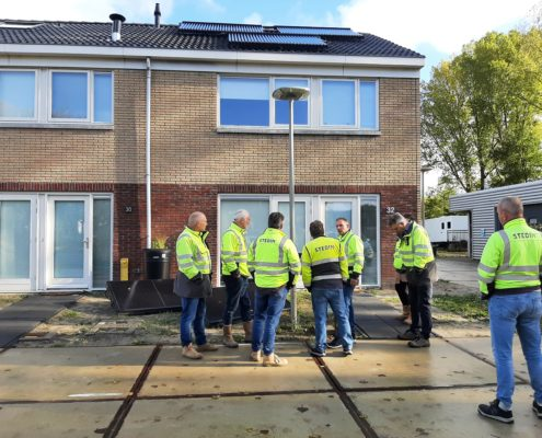 Een groep mensen in felgele veiligheidsjassen staat op een stoep voor een bakstenen woonhuis met zonnepanelen op het dak.