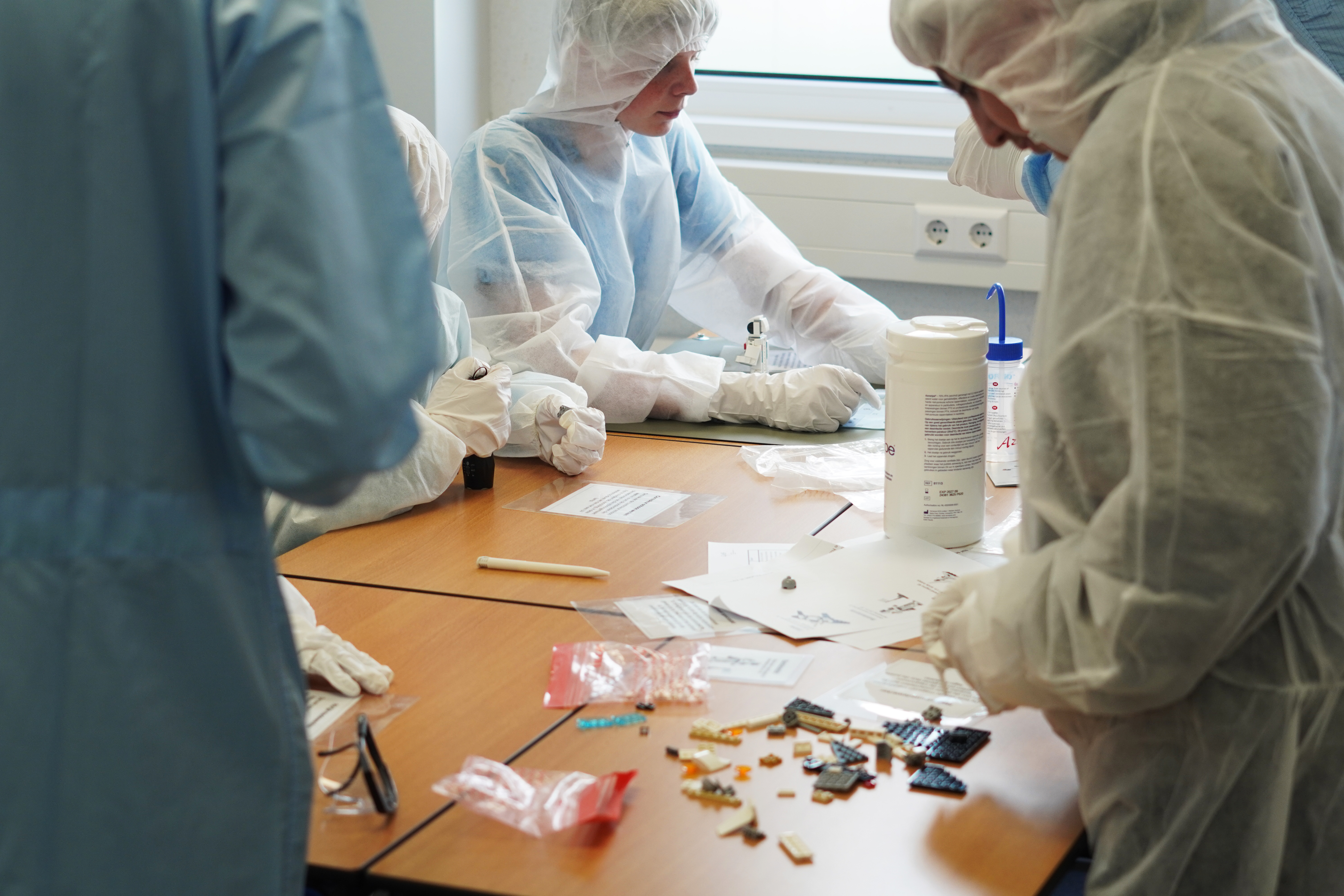 Personen in beschermende kleding werken samen aan een praktische oefening aan een tafel met kleine onderdelen en materialen