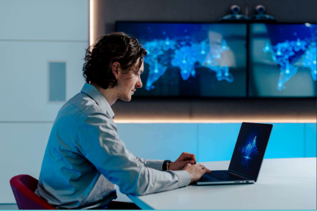 Een persoon werkt achter een laptop in een moderne kantoorruimte, met op de achtergrond schermen waarop een digitale wereldkaart zichtbaar is.