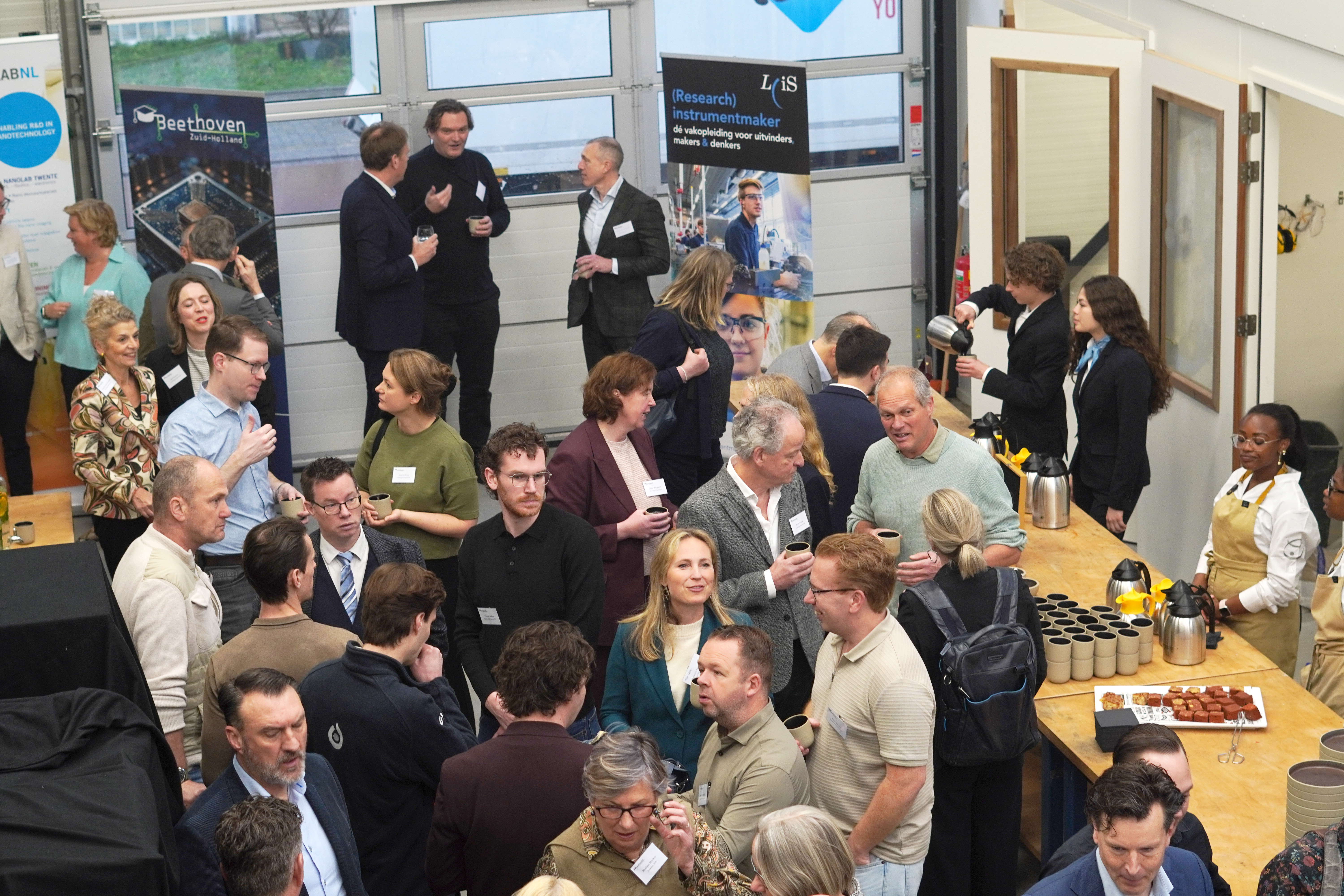 Bezoekers praten en netwerken tijdens een druk evenement, met koffietafel en informatiestands op de achtergrond.