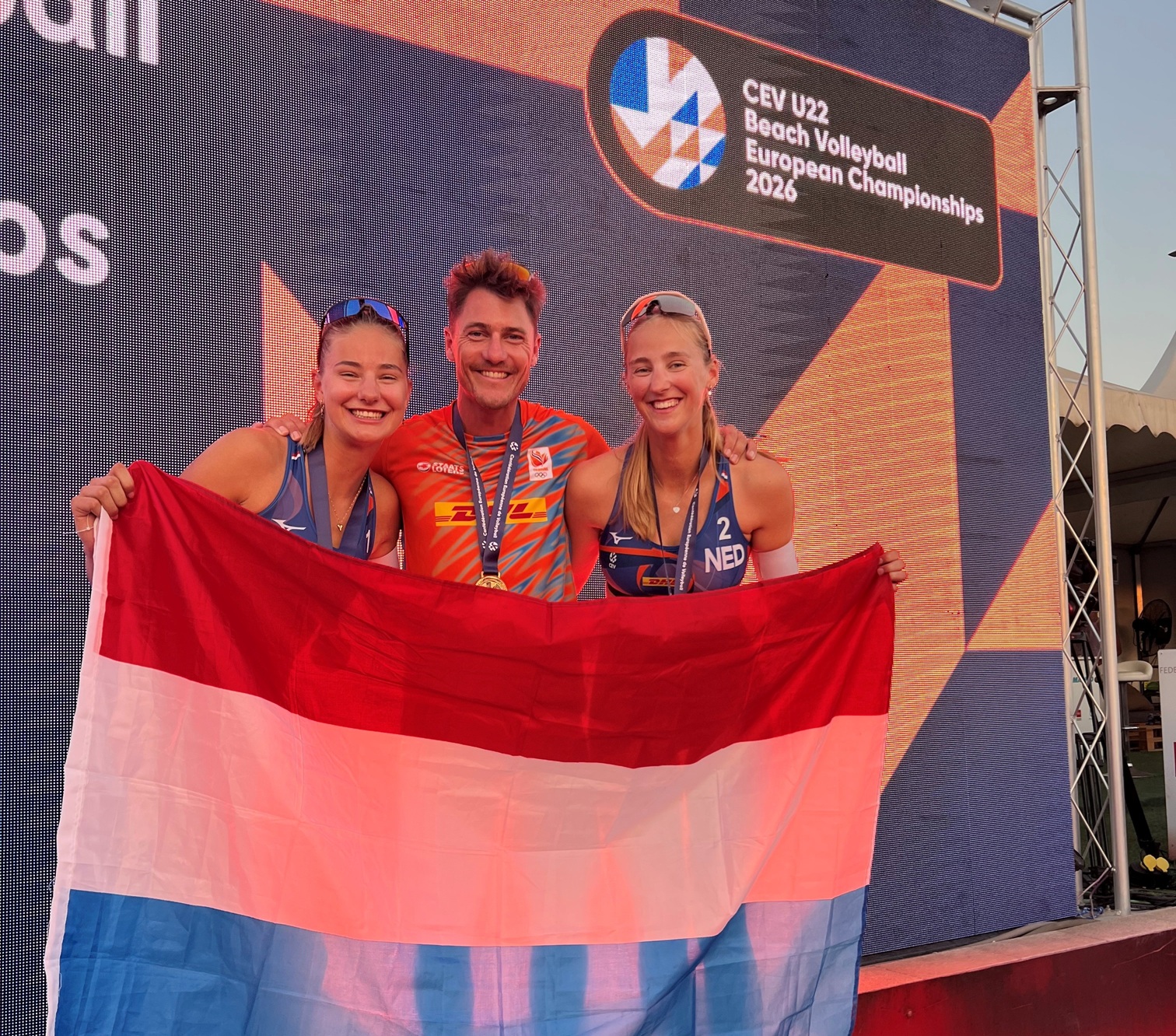 Drie personen poseren met de Nederlandse vlag tijdens het EK beachvolleybal onder 22 in 2026.