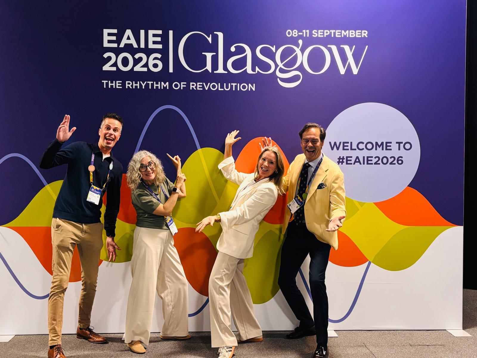 Vier deelnemers poseren voor de kleurrijke EAIE 2026 Glasgow-fotowand met de tekst “The Rhythm of Revolution